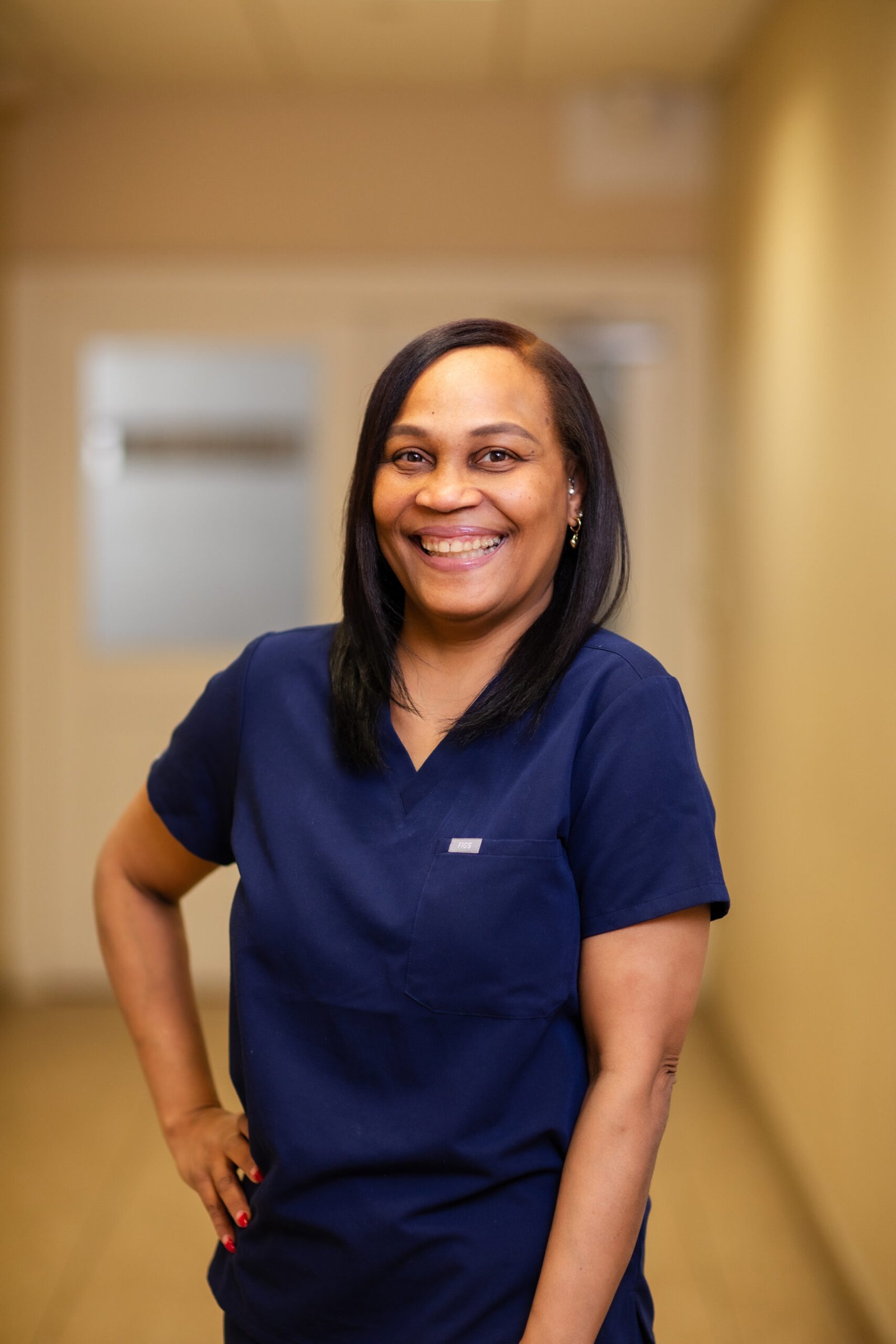 Smiling woman with straight dark hair, wearing navy scrubs, standing with hand on hip for a staff portrait.