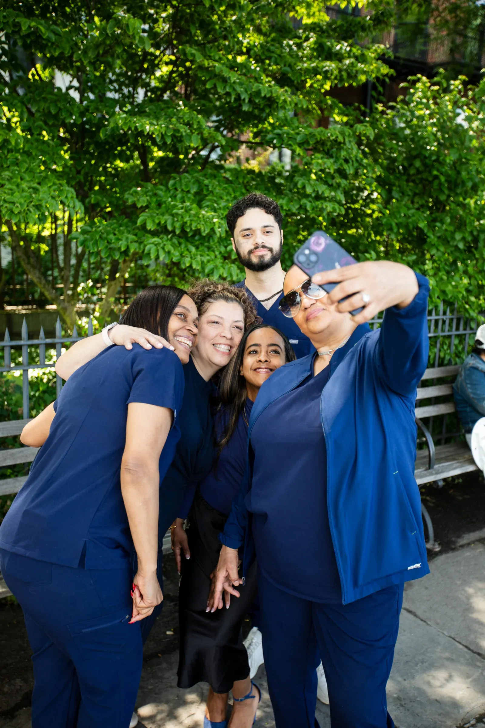 Dental office team in navy scrubs smiling and taking a group selfie outdoors in front of green trees