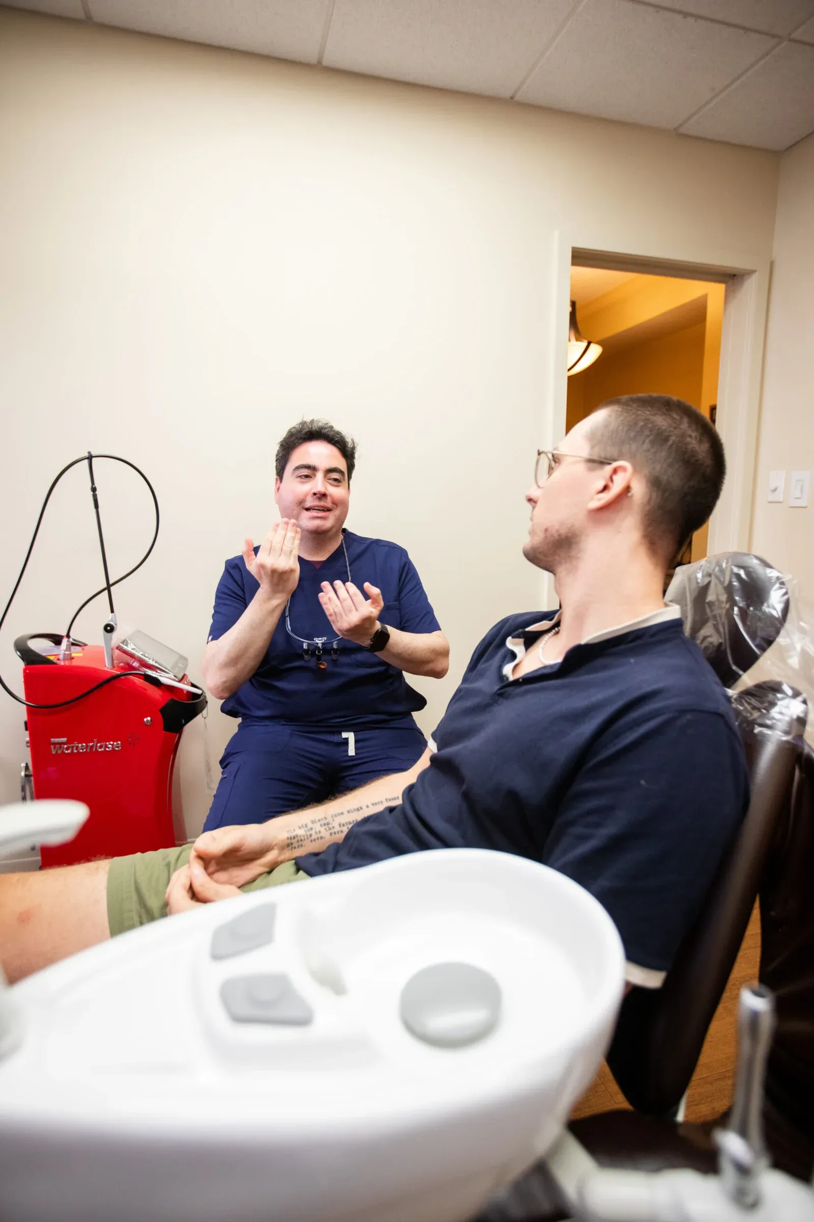 Dentist in navy scrubs gesturing while explaining treatment to a seated patient, with a Waterlase laser unit visible in the background