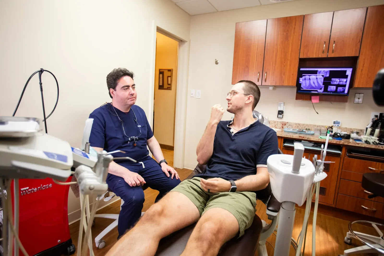 Dentist in navy scrubs talking with a seated male patient in the dental chair, with dental X-ray images displayed on a wall-mounted monitor in the background.