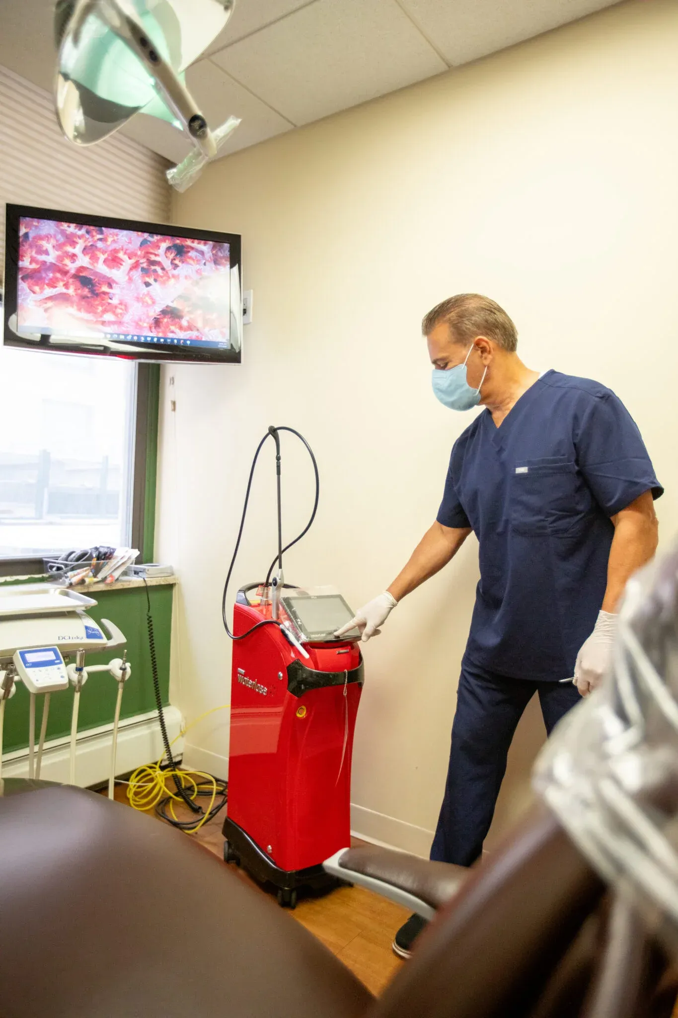 Dentist wearing a mask adjusts the touchscreen controls on a red Waterlase dental laser unit, with an intraoral camera view of gum tissue shown on the wall monitor behind him.