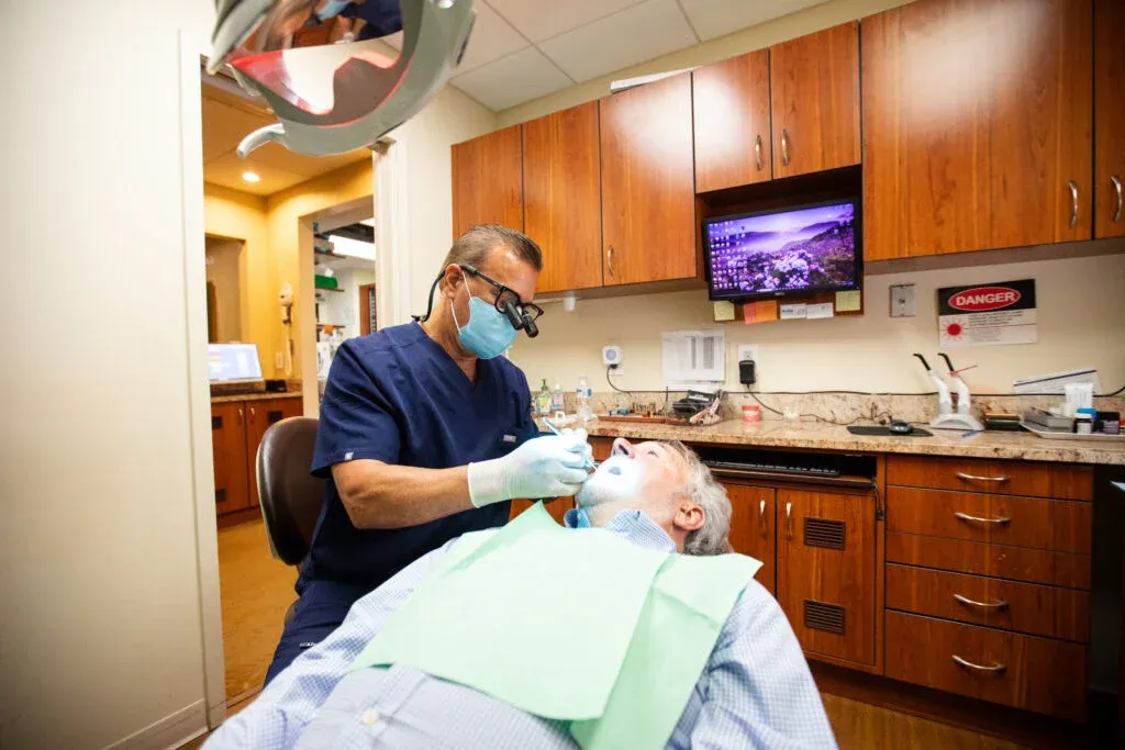 Dentist wearing a mask and magnifying loupes performs a procedure on a reclined male patient's teeth in a wood-cabinetry treatment room, with a laser device and monitor visible nearby.