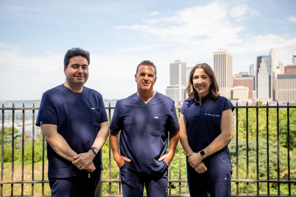 Dr. Alexander Black, Dr. Matthew Bellafiore, and Dr. Rebecca Baer standing together with the Manhattan skyline in the background, dentists at Creative Dentistry of Brooklyn Heights