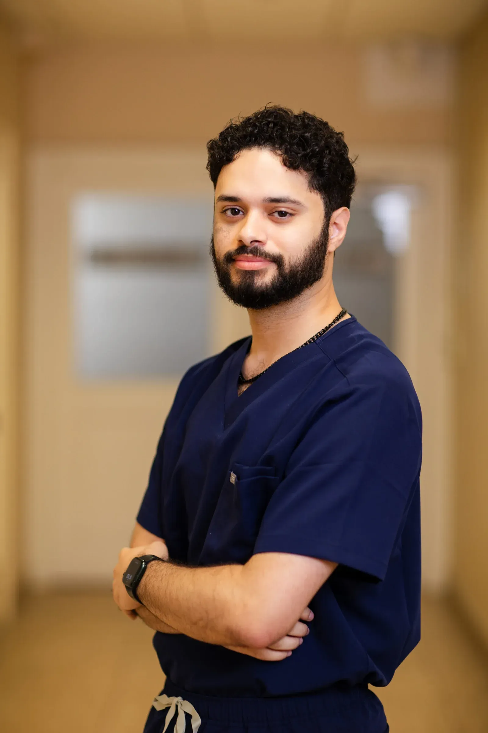 Man with curly dark hair and a beard, wearing navy scrubs, standing with arms crossed for a staff portrait.