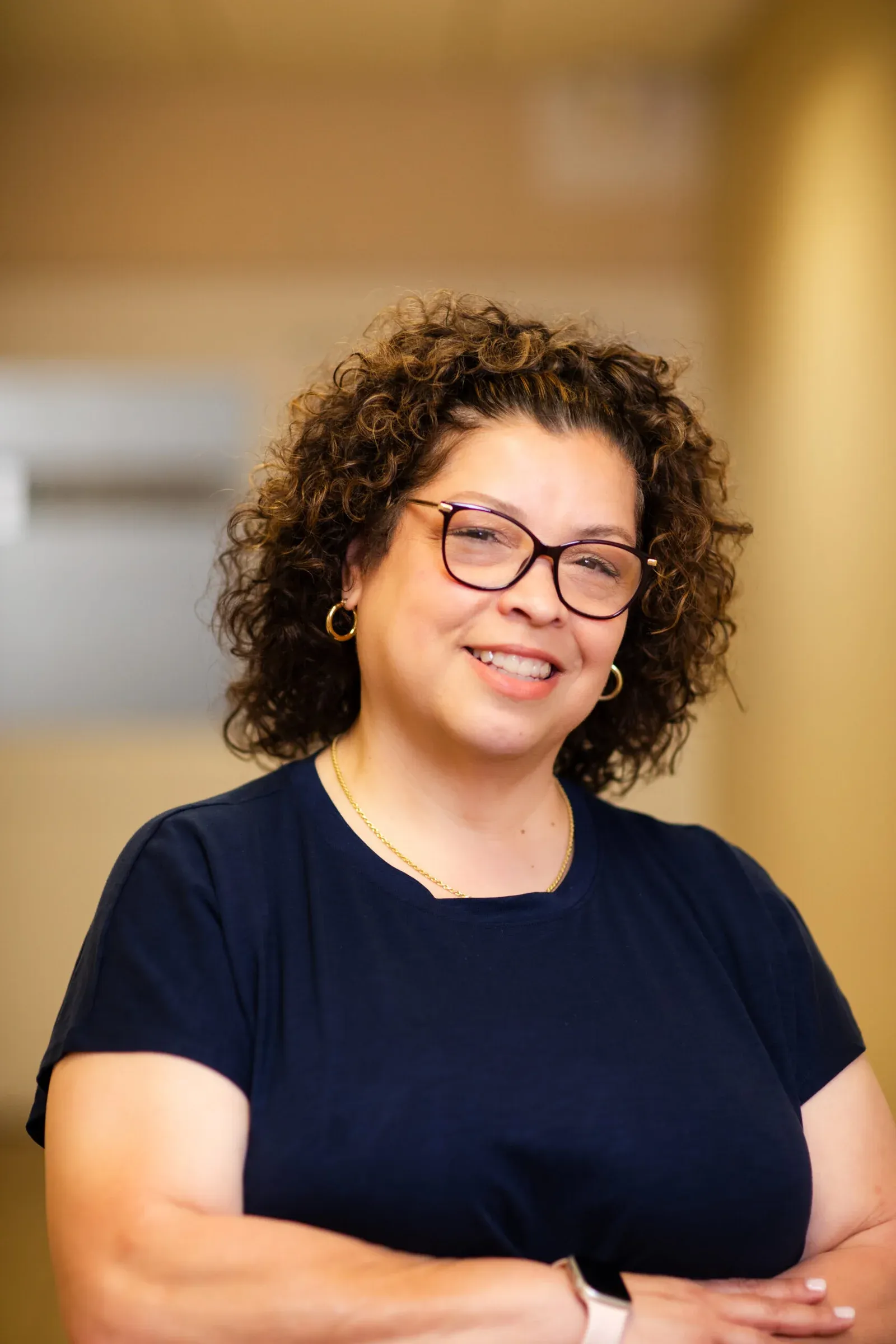 Smiling woman with curly hair and glasses, wearing a navy top, posing for a staff portrait.