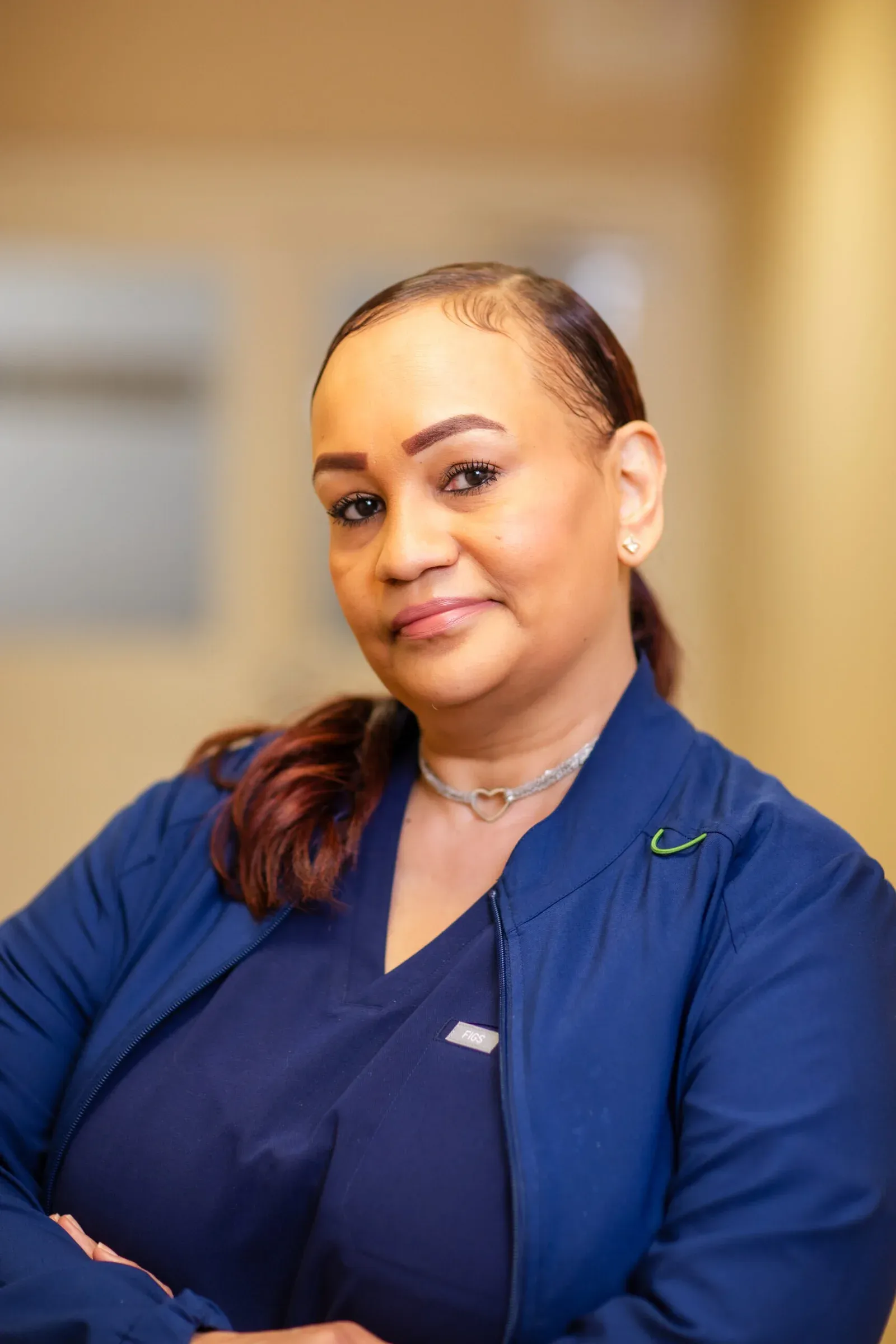 Woman with hair pulled back, wearing a navy scrub jacket and heart-pendant necklace, posing for a staff portrait.