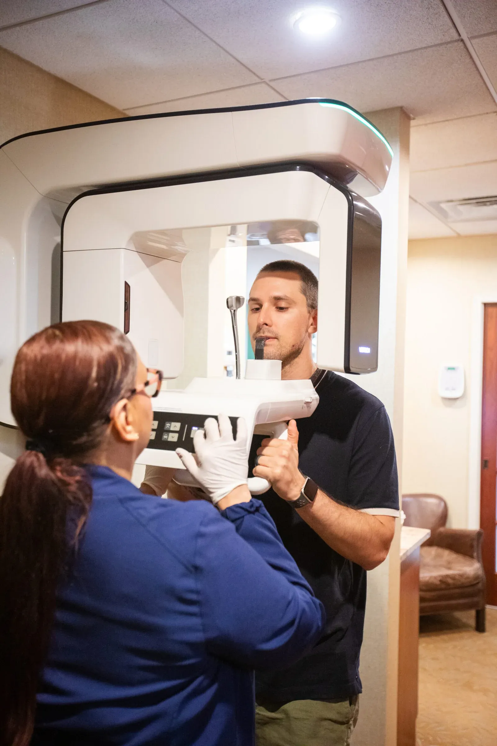 Dental assistant in blue scrubs and gloves positioning a male patient in a panoramic dental X-ray machine, patient biting down on the positioning guide.