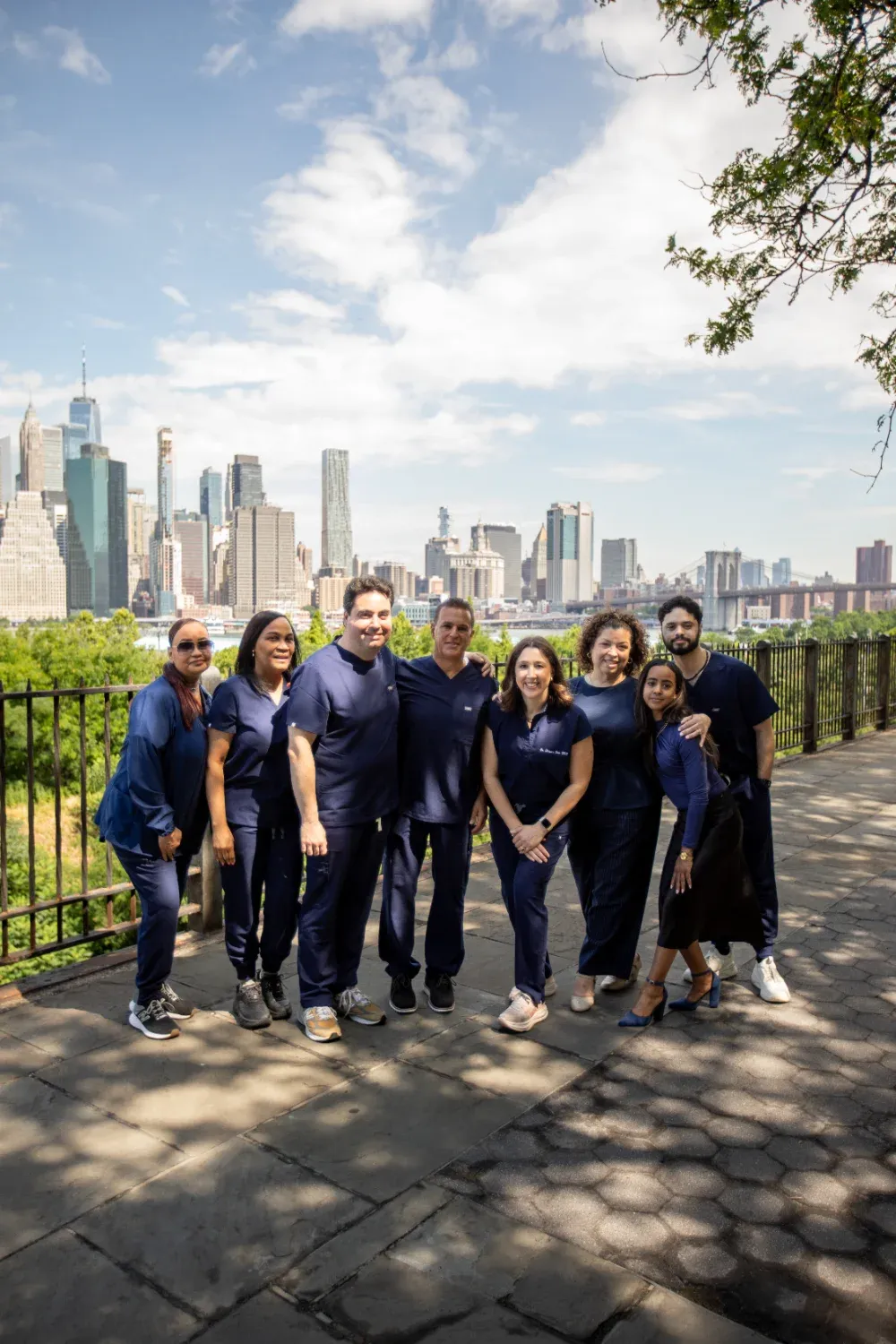 Full team photo of Creative Dentistry of Brooklyn Heights staff, including Dr. Alexander Black, Dr. Matthew Bellafiore, and Dr. Rebecca Baer, at the Brooklyn Heights Promenade with the Manhattan skyline and Brooklyn Bridge in the background