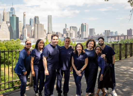Full dental office team in navy scrubs posing together on a waterfront promenade, with the Manhattan skyline and Brooklyn Bridge in the background