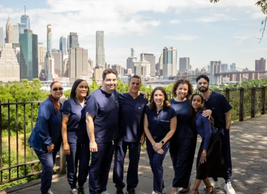 Full team photo of Creative Dentistry of Brooklyn Heights staff, including Dr. Alexander Black, Dr. Matthew Bellafiore, and Dr. Rebecca Baer, at the Brooklyn Heights Promenade with the Manhattan skyline and Brooklyn Bridge in the background